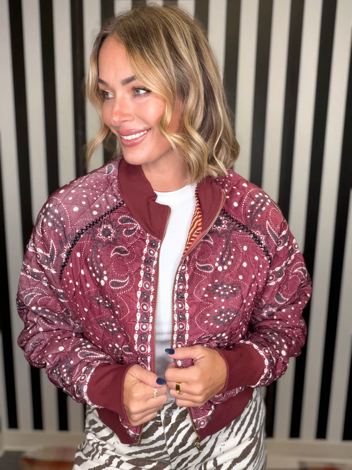 A woman wearing a burgundy and white paisley bandana bomber jacket over a white t-shirt, standing in front of a black and white striped wall.