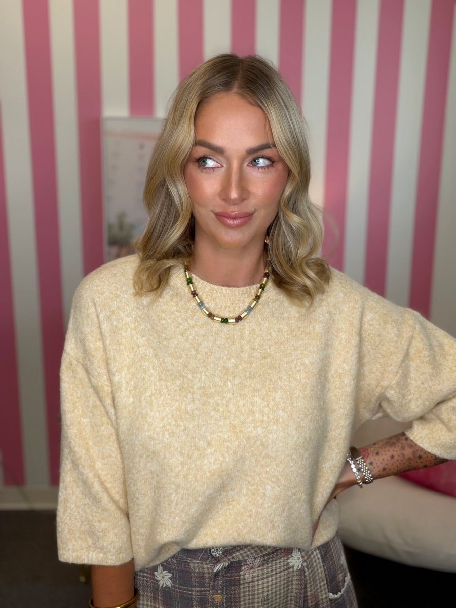 A woman with blonde wavy hair wearing a cream-colored, textured knit crewneck sweater and a beaded necklace, standing in front of a pink and white striped wall.