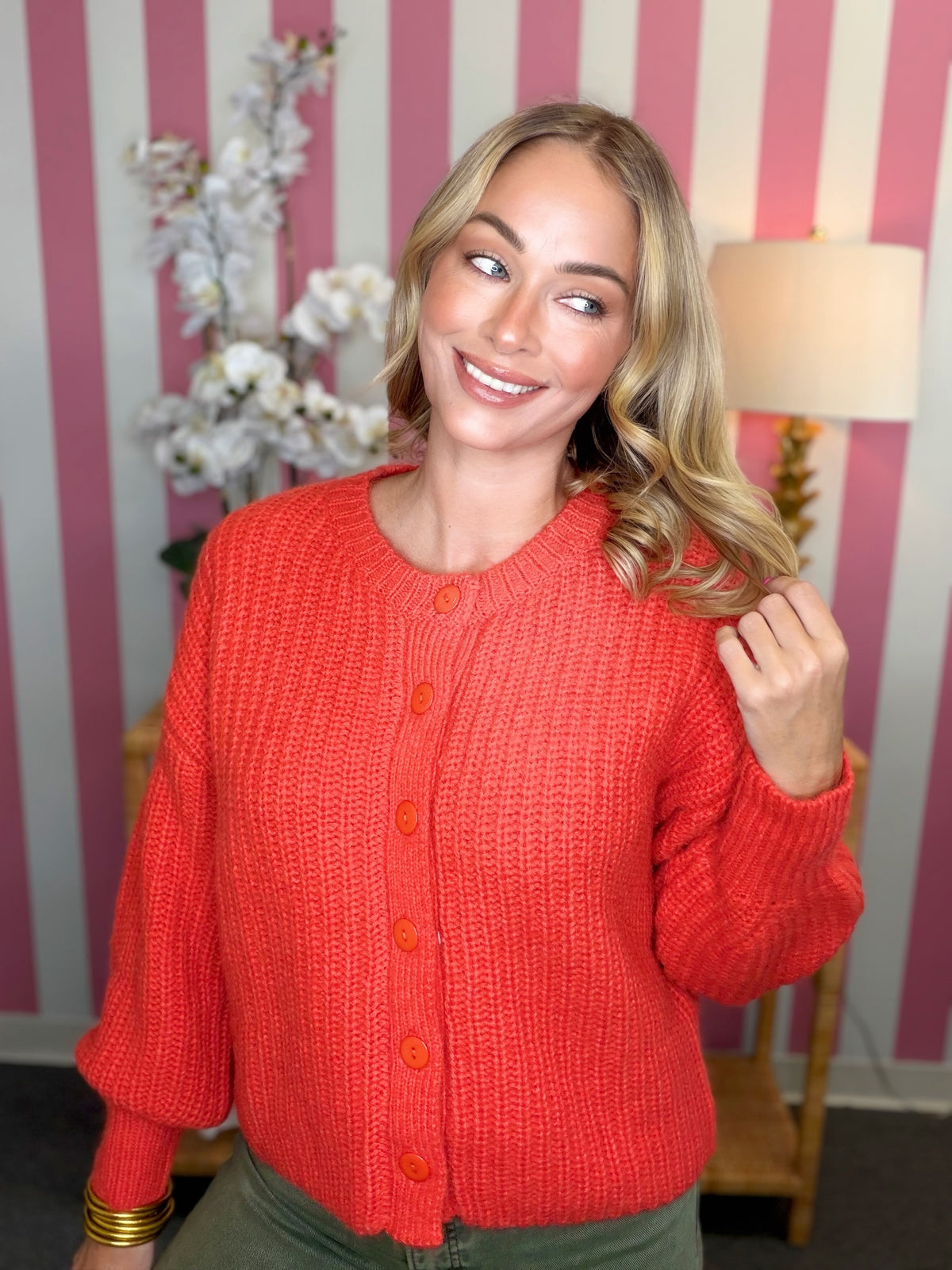 A woman wearing a bright coral-red textured knit cardigan sweater with button-front closure, standing in front of a pink and white striped wall with a lamp and flowers in the background.