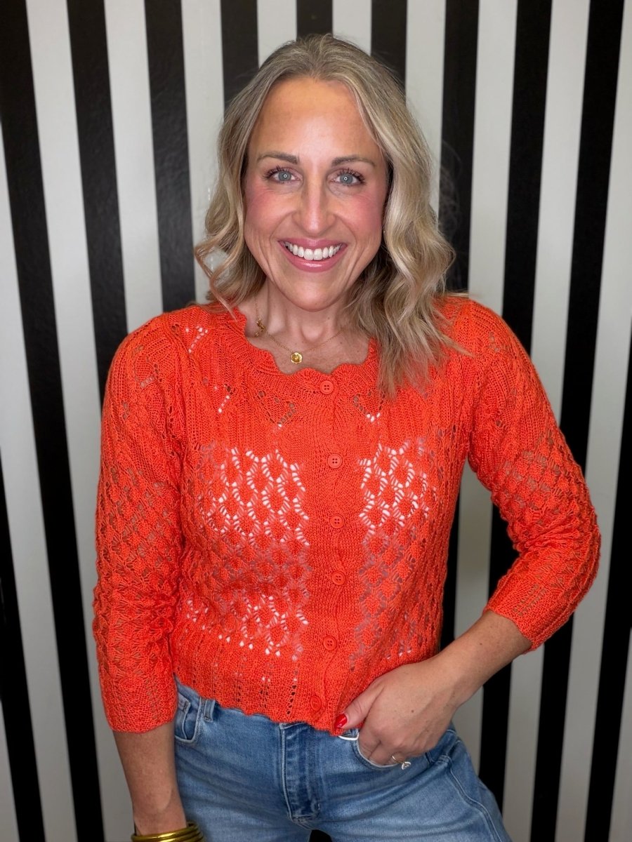 A smiling blonde woman wearing a bright orange open-knit long-sleeve top and blue jeans, standing against a black and white vertically striped background.