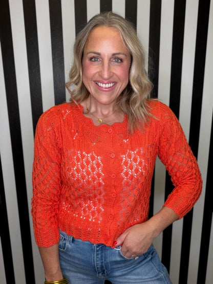 A smiling blonde woman wearing a bright orange open-knit long-sleeve top and blue jeans, standing against a black and white vertically striped background.