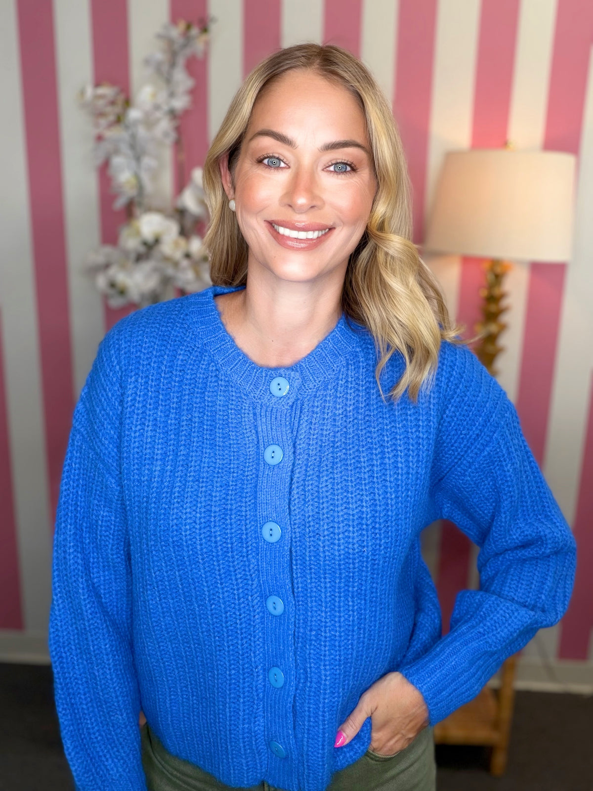 A smiling blonde woman wearing a bright blue textured knit cardigan sweater with matching buttons, standing in front of a pink and white striped wall with a lamp in the background.