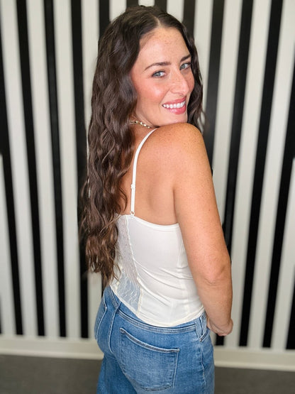 A woman with long wavy brown hair is shown from the back wearing a white spaghetti-strap camisole top and blue jeans, standing in front of a black and white vertically striped wall.