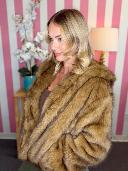 A woman with blonde hair wearing a plush brown faux fur jacket, standing in front of a pink and white striped wall with a lamp and flowers in the background.