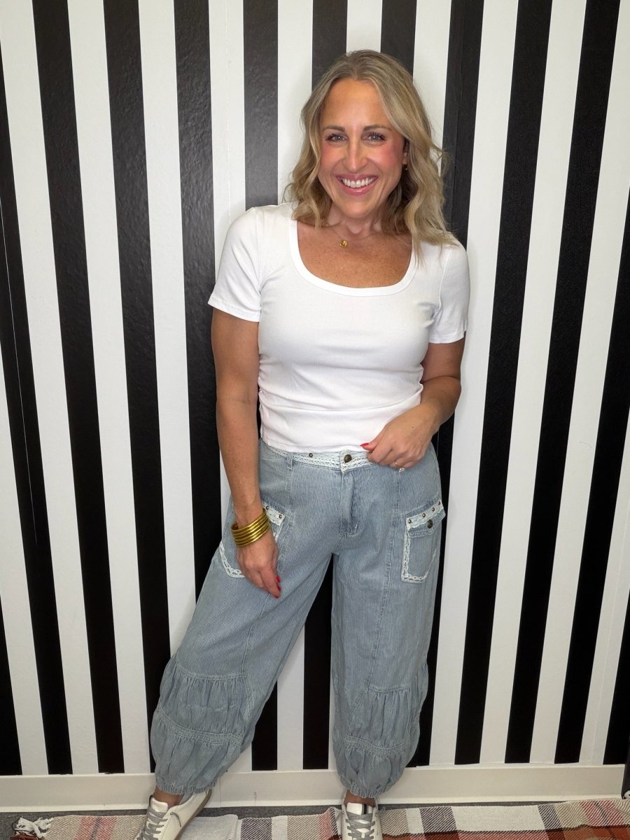 A woman wearing a white t-shirt and light wash denim baggy pants with ruffle details, standing in front of a black and white striped wall.