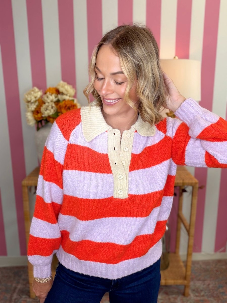 A woman wearing a knit polo-style sweater with bold horizontal red and light lavender stripes, a cream ribbed collar, and a cream button placket, standing in front of pink and white striped wallpaper.