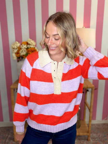 A woman wearing a knit polo-style sweater with bold horizontal red and light lavender stripes, a cream ribbed collar, and a cream button placket, standing in front of pink and white striped wallpaper.