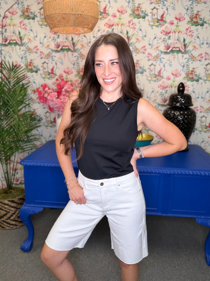 Woman in black sleeveless top and white shorts standing in a room with floral wallpaper and blue furniture.