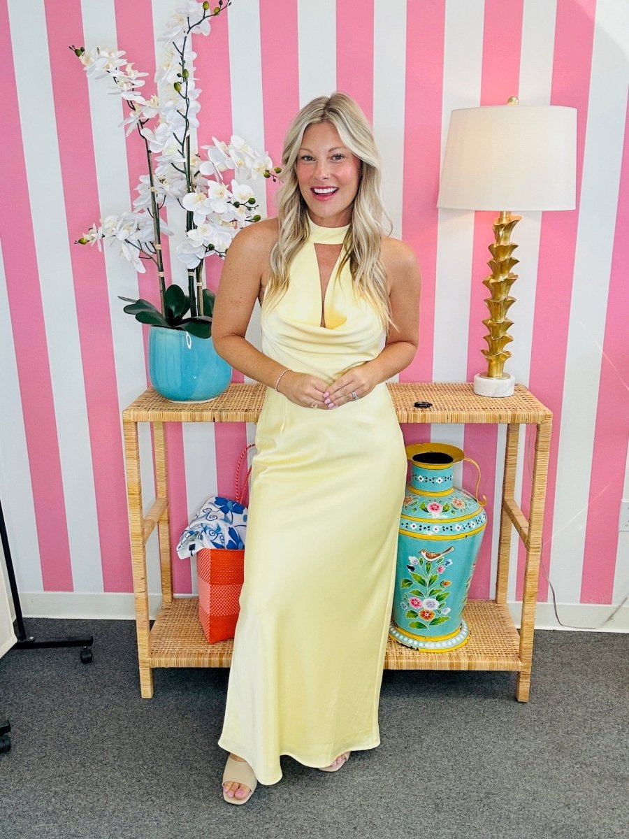 Woman in a yellow dress standing in front of a pink and white striped wall with decorative items on a table.