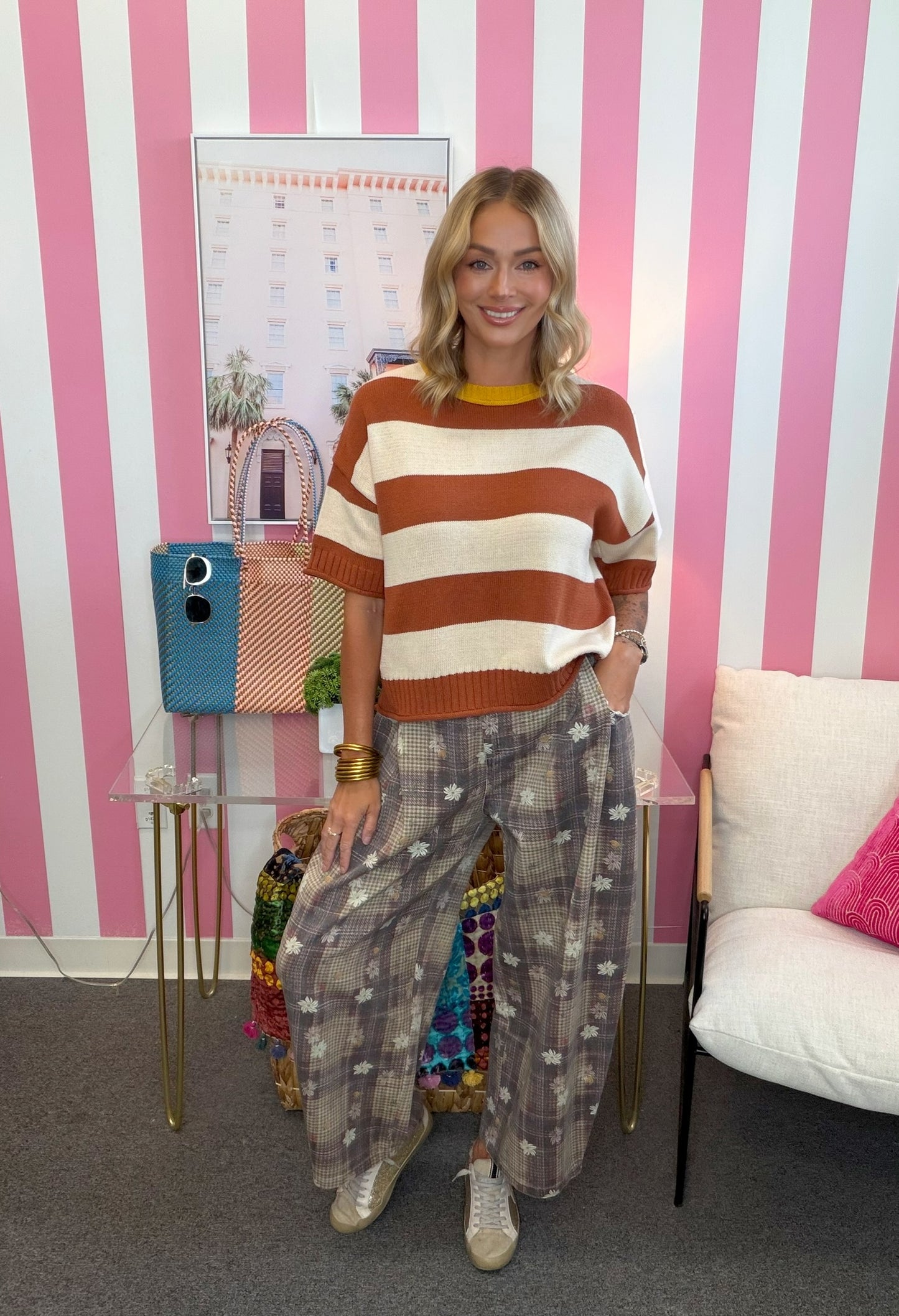 A woman stands in a room with pink and white vertical stripes, wearing a rust orange and cream striped knit sweater, patterned wide-leg trousers, and beige sneakers.
