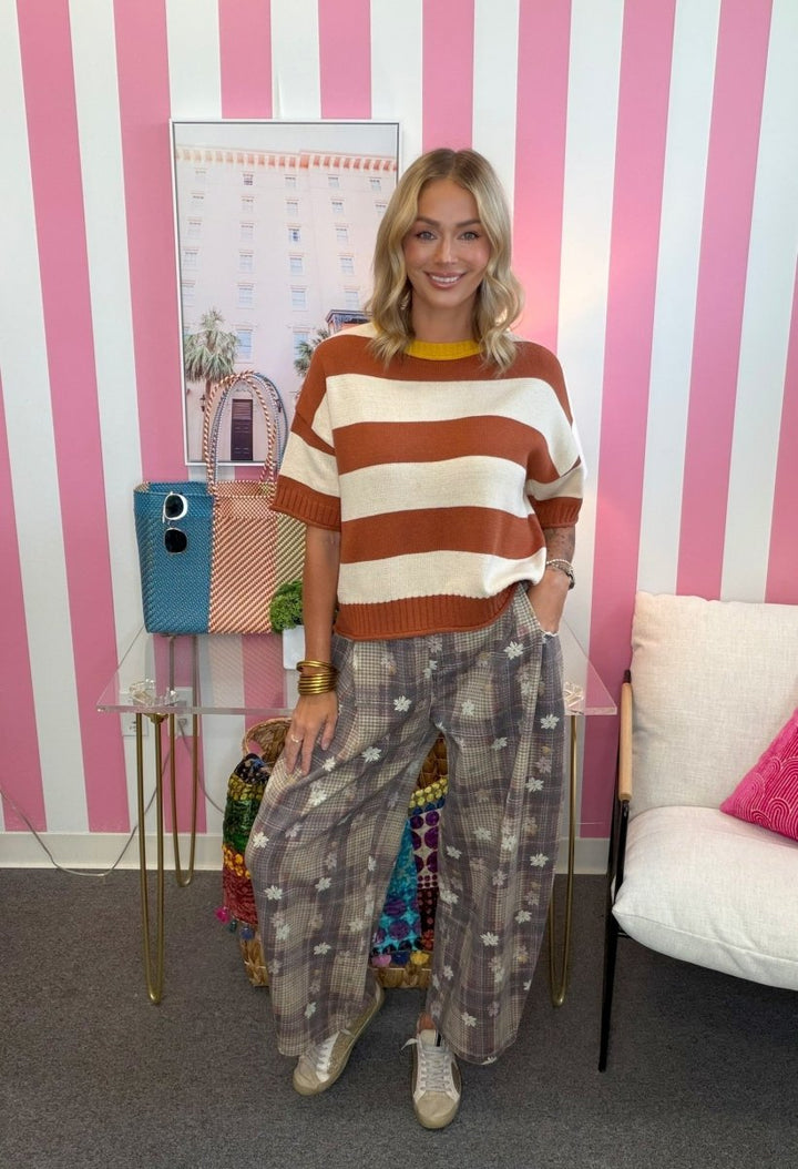 A woman stands in a room with pink and white vertical stripes, wearing a rust orange and cream striped knit sweater, patterned wide-leg trousers, and beige sneakers.