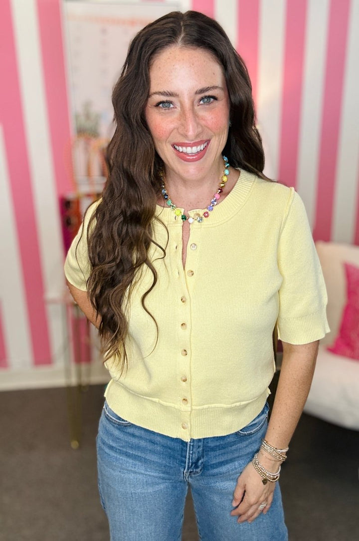 Woman wearing a yellow shirt and blue jeans with a colorful necklace against a pink and white striped background.