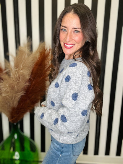 A smiling woman with long brown hair wearing a light gray knit sweater with blue polka dots and light blue jeans, standing in front of a black and white striped wall with a green glass vase and dried grass in the foreground.