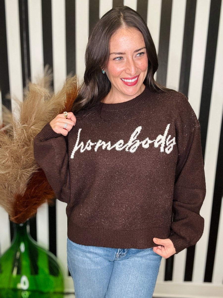 A woman wearing a brown knit sweater with 'homebody' written across the chest in white script, paired with light blue jeans, standing in front of a black and white striped background.
