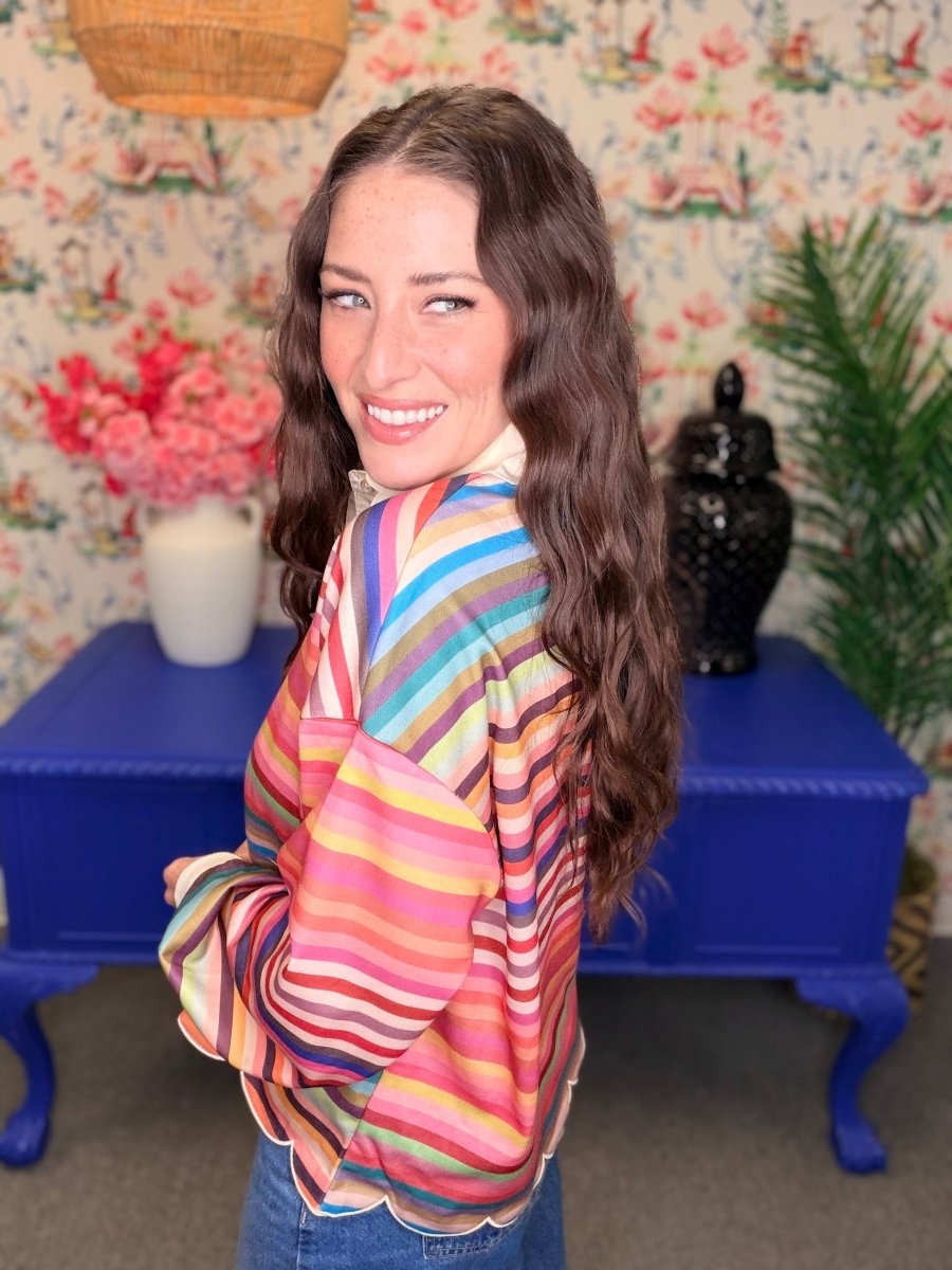 A woman with long wavy brown hair smiles while wearing a colorful striped long-sleeve blouse with a scalloped hem, standing in a decorated room with floral wallpaper, a blue side table, and plants.