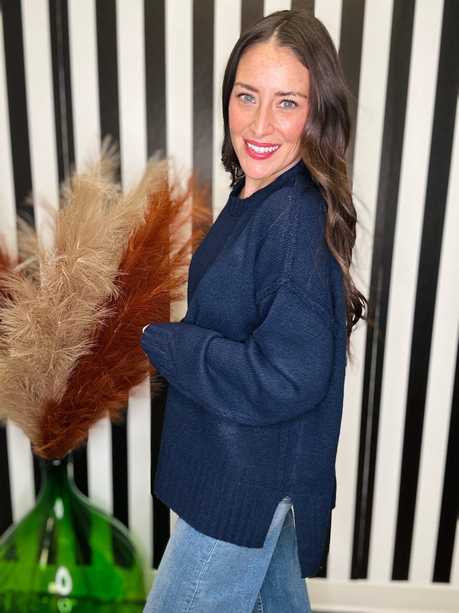 A woman wearing a navy blue knit sweater and light blue jeans, standing in front of a black and white striped wall with a green glass vase and dried pampas grass on the left.