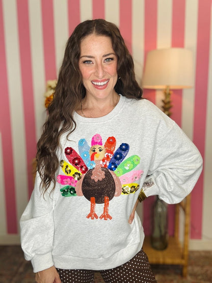 A woman wearing a light gray crewneck sweatshirt with a colorful sequined turkey graphic on the front, posed in front of a pink and white striped wall.