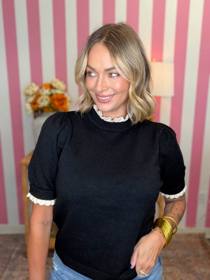 A woman wearing a black short-sleeve knit sweater with white ruffled trim at the collar and sleeve cuffs, paired with light blue jeans. She is standing in front of a pink and white striped wall.