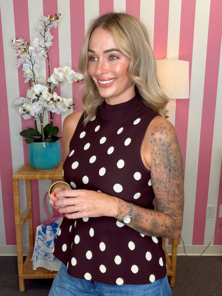 A woman with blonde hair and tattoos wearing a sleeveless mock neck top with white polka dots on a dark burgundy background, standing in front of pink and white striped wallpaper.