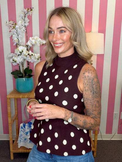 A woman with blonde hair and tattoos wearing a sleeveless mock neck top with white polka dots on a dark burgundy background, standing in front of pink and white striped wallpaper.