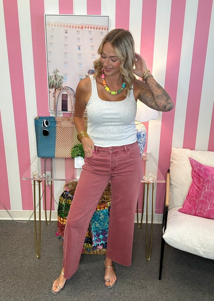 A woman modeling dusty rose wide-leg jeans and a white ribbed tank top in a pink and white striped room, accessorized with a colorful beaded necklace and gold bracelets.