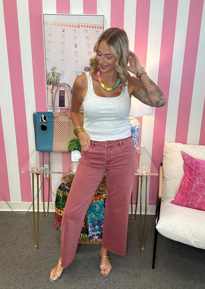 A woman modeling dusty rose wide-leg jeans and a white ribbed tank top in a pink and white striped room, accessorized with a colorful beaded necklace and gold bracelets.
