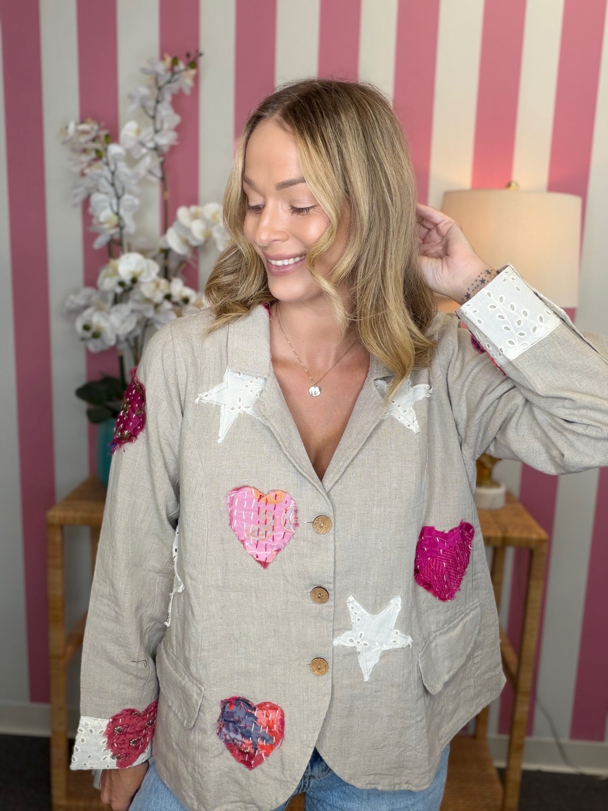 A woman wearing a beige linen-blend blazer decorated with white star appliqués and pink, red, and blue heart appliqués, posed in front of a pink and white striped wall.