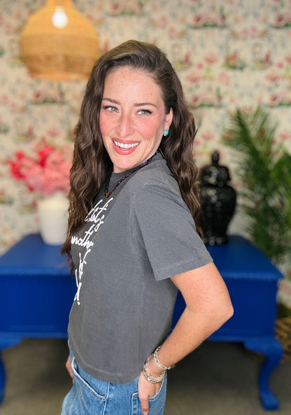 A woman with long wavy brown hair smiling and looking over her shoulder, wearing a dark gray graphic t-shirt and blue jeans, standing in a room with floral wallpaper, a blue table, and a wicker pendant light.