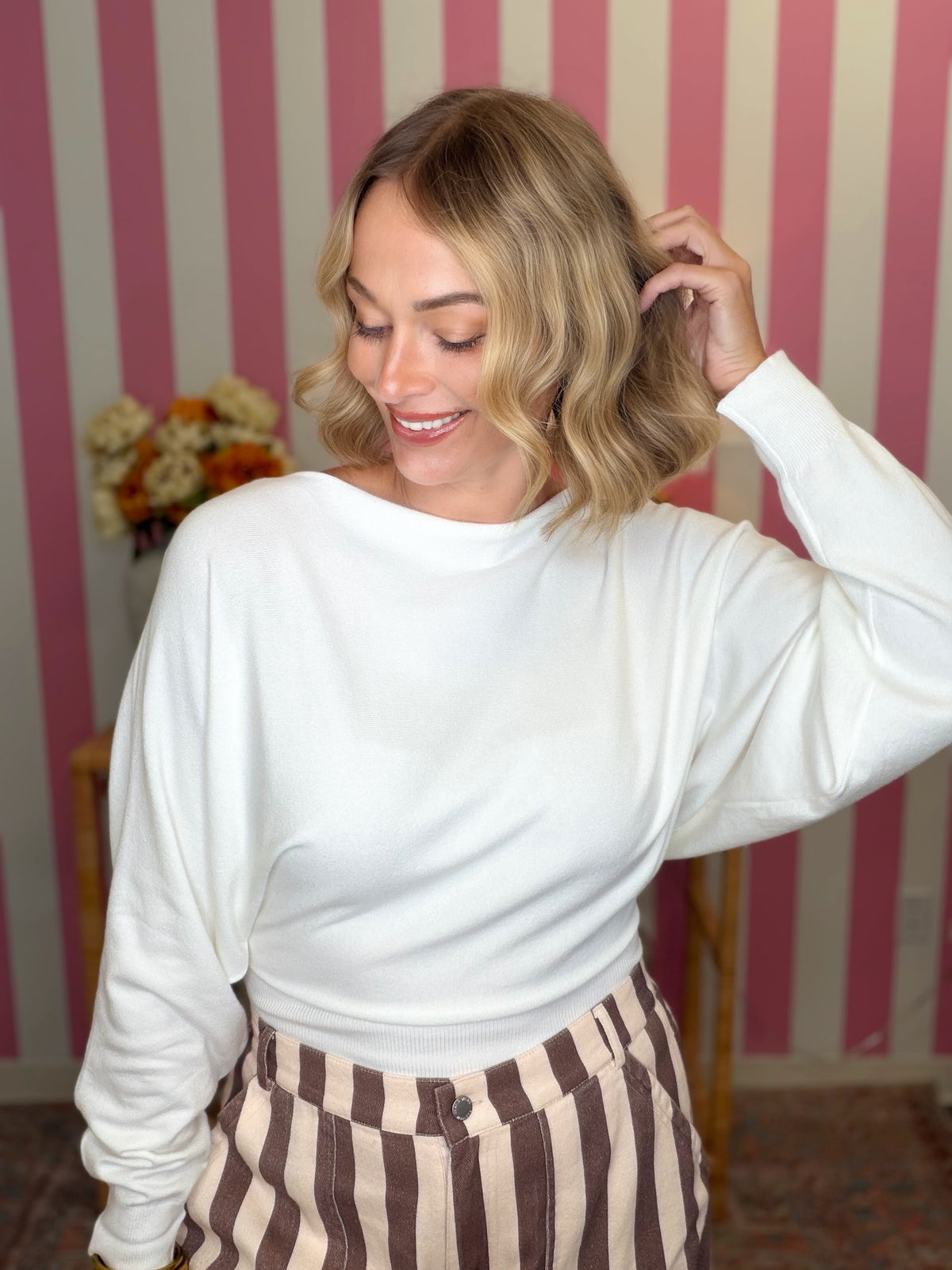 A woman with blonde wavy hair wearing a white long-sleeve crewneck top and brown and beige striped pants, posing in front of a pink and white striped wall.