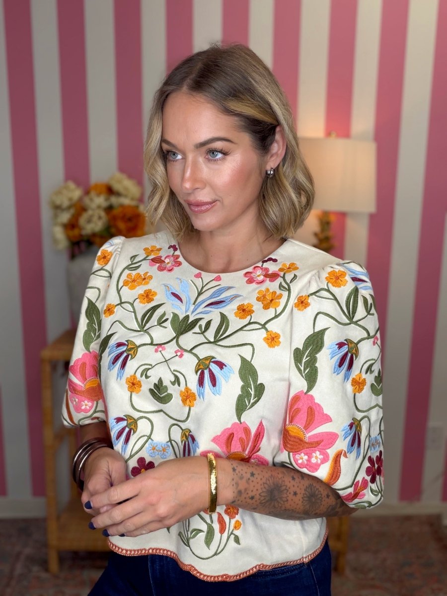 A woman wearing a cream-colored short-sleeve blouse with a vibrant multicolor floral and butterfly print, paired with dark blue jeans, standing in front of a pink and white striped wall.