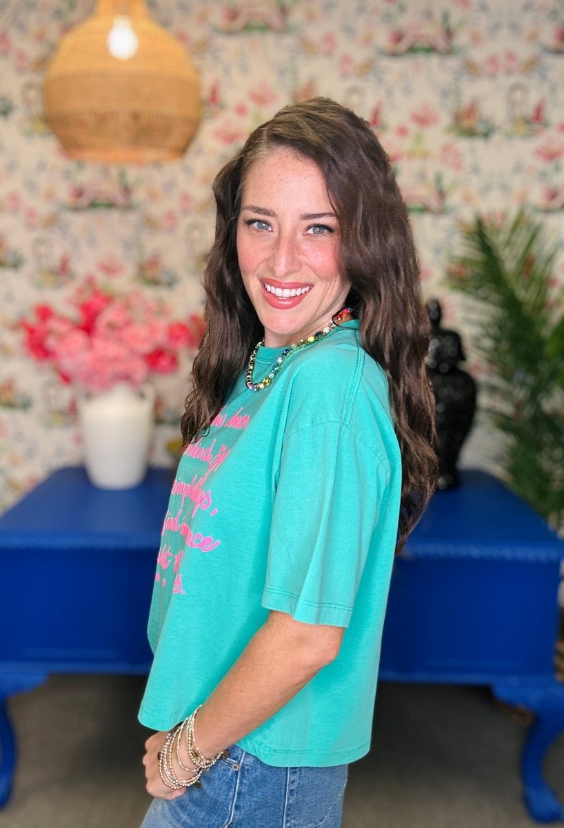 A woman with long brown hair smiling and looking over her shoulder, wearing a turquoise short-sleeve graphic t-shirt with pink text and a green beaded necklace, standing in a room with floral wallpaper, a blue table, and a hanging lamp.