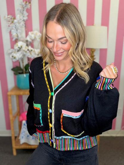 A woman wearing a black knit cardigan with vibrant multicolor striped trim and colorful embroidered pockets, posed in front of a pink and white striped wall.