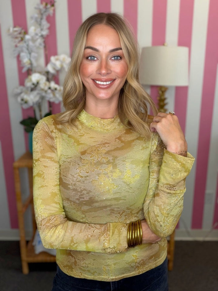 A smiling blonde woman wearing a long-sleeve yellow and gold patterned mock neck top, standing in front of a pink and white striped wall.
