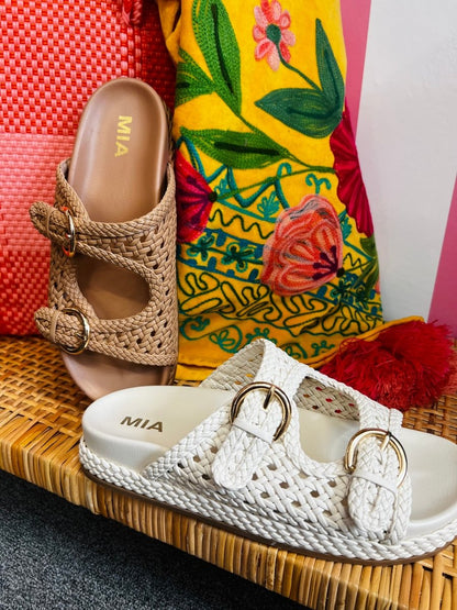 Pair of woven sandals with gold buckles on a textured surface with colorful fabric in the background.