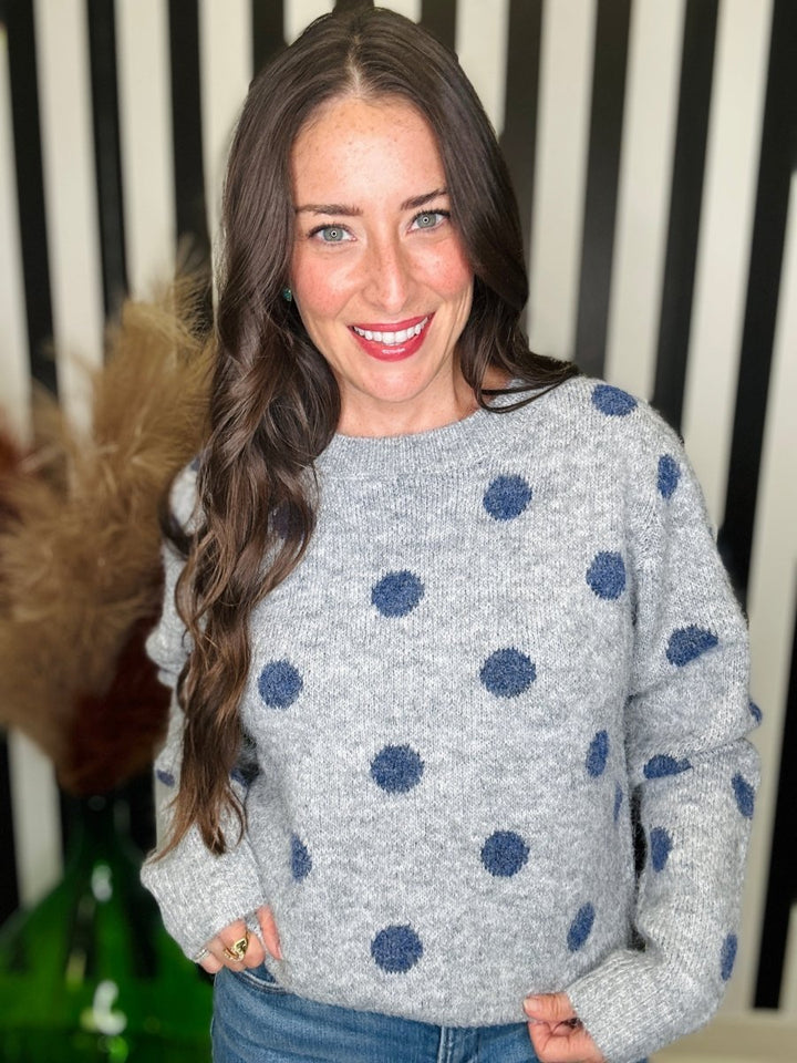 A smiling woman with long brown hair wearing a light gray knit sweater with dark blue polka dots, paired with blue jeans, standing in front of a black and white striped background.