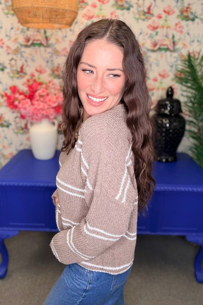 A woman with long wavy brown hair smiles while looking over her shoulder, wearing a chunky knit cropped cardigan in taupe with white stripes and blue jeans. She is standing in front of a blue table against a floral wallpaper background.