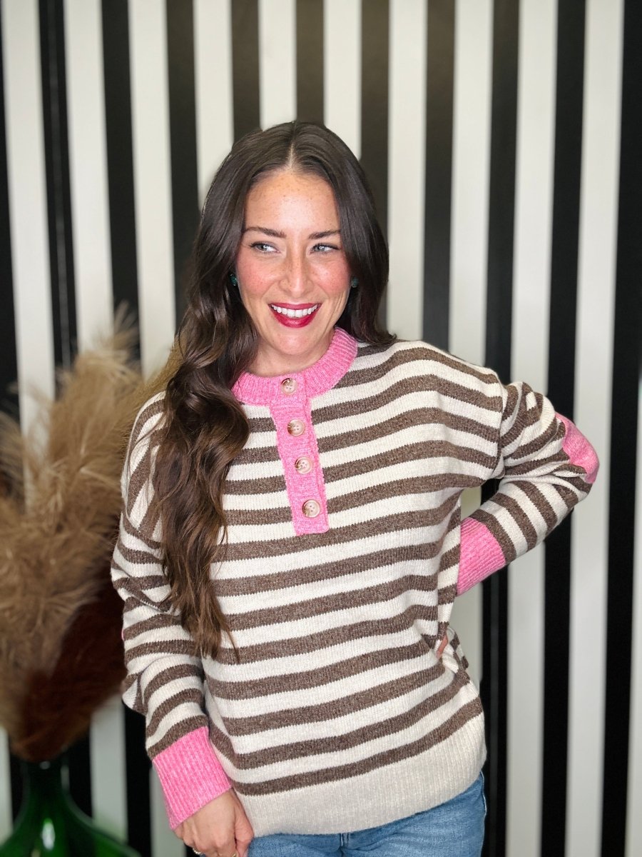 A woman wearing a brown and cream striped knit sweater with bright pink trim on the henley placket and cuffs, posed in front of a black and white striped background.