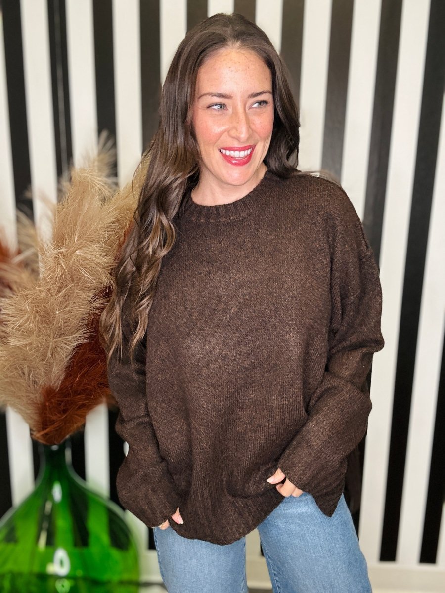 A woman wearing a brown knit crewneck sweater and light blue jeans, standing in front of a black and white striped wall with a green glass vase and dried grass on the left.