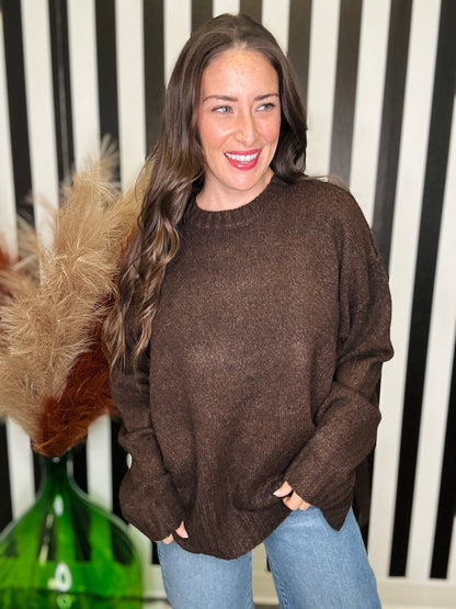A woman wearing a brown knit crewneck sweater and light blue jeans, standing in front of a black and white striped wall with a green glass vase and dried grass on the left.