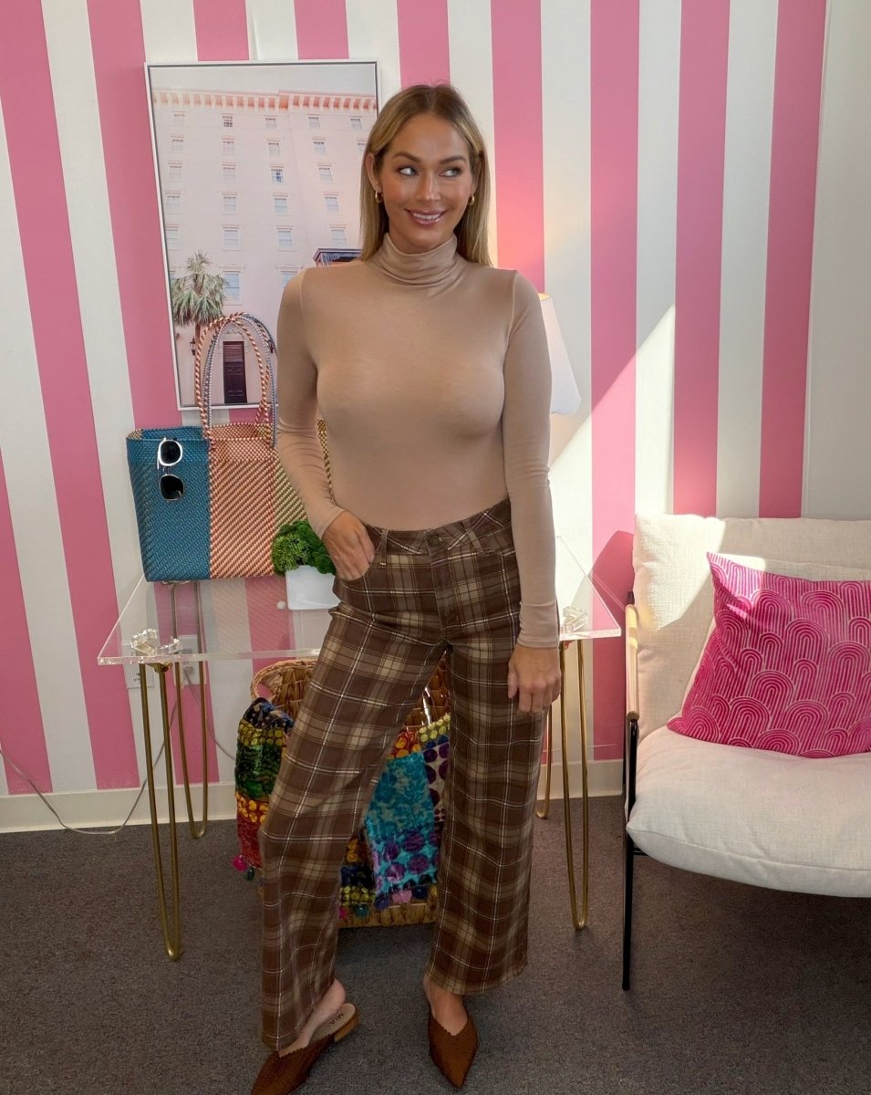 A woman stands in a room with pink and white striped walls, wearing a beige mock-neck long-sleeve top and brown plaid wide-leg trousers.