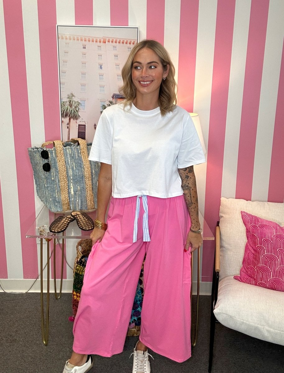 A woman stands in a room with pink and white striped wallpaper, wearing a white short-sleeve t-shirt and bright pink wide-leg pants with a white drawstring waist.