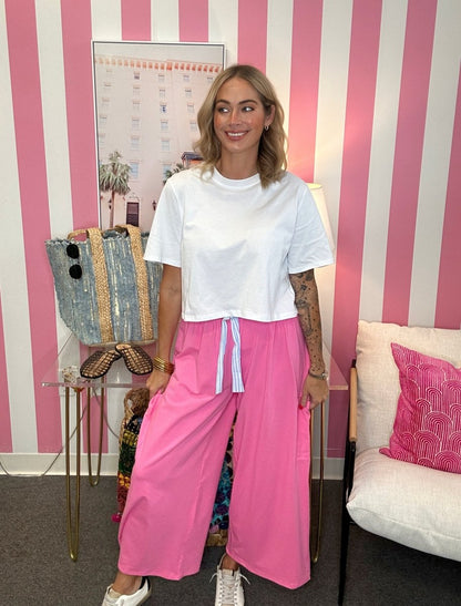 A woman stands in a room with pink and white striped wallpaper, wearing a white short-sleeve t-shirt and bright pink wide-leg pants with a white drawstring waist.