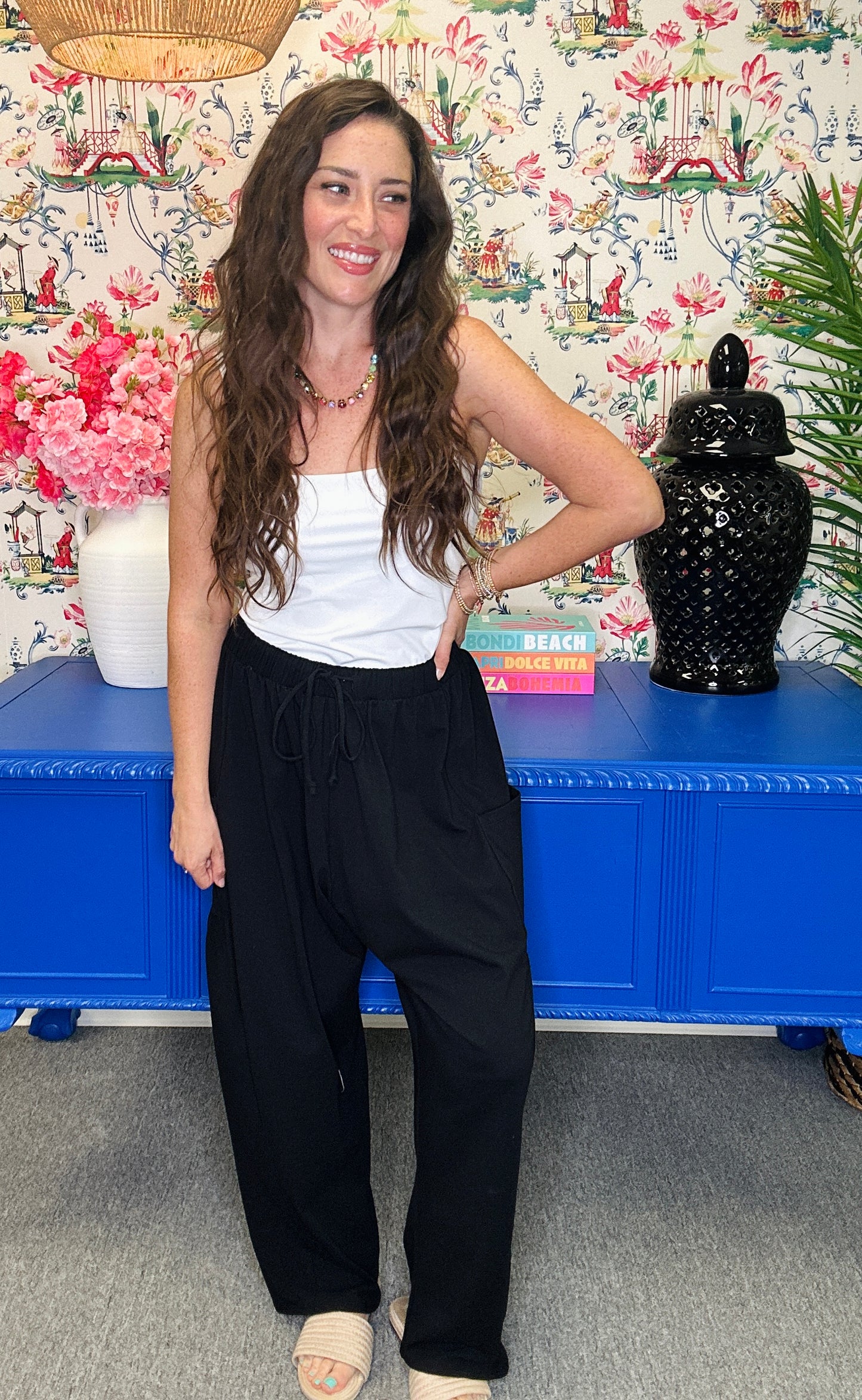 A woman posing in a white ribbed tank top and black wide-leg trousers, standing in front of a blue cabinet and a floral wallpaper background.