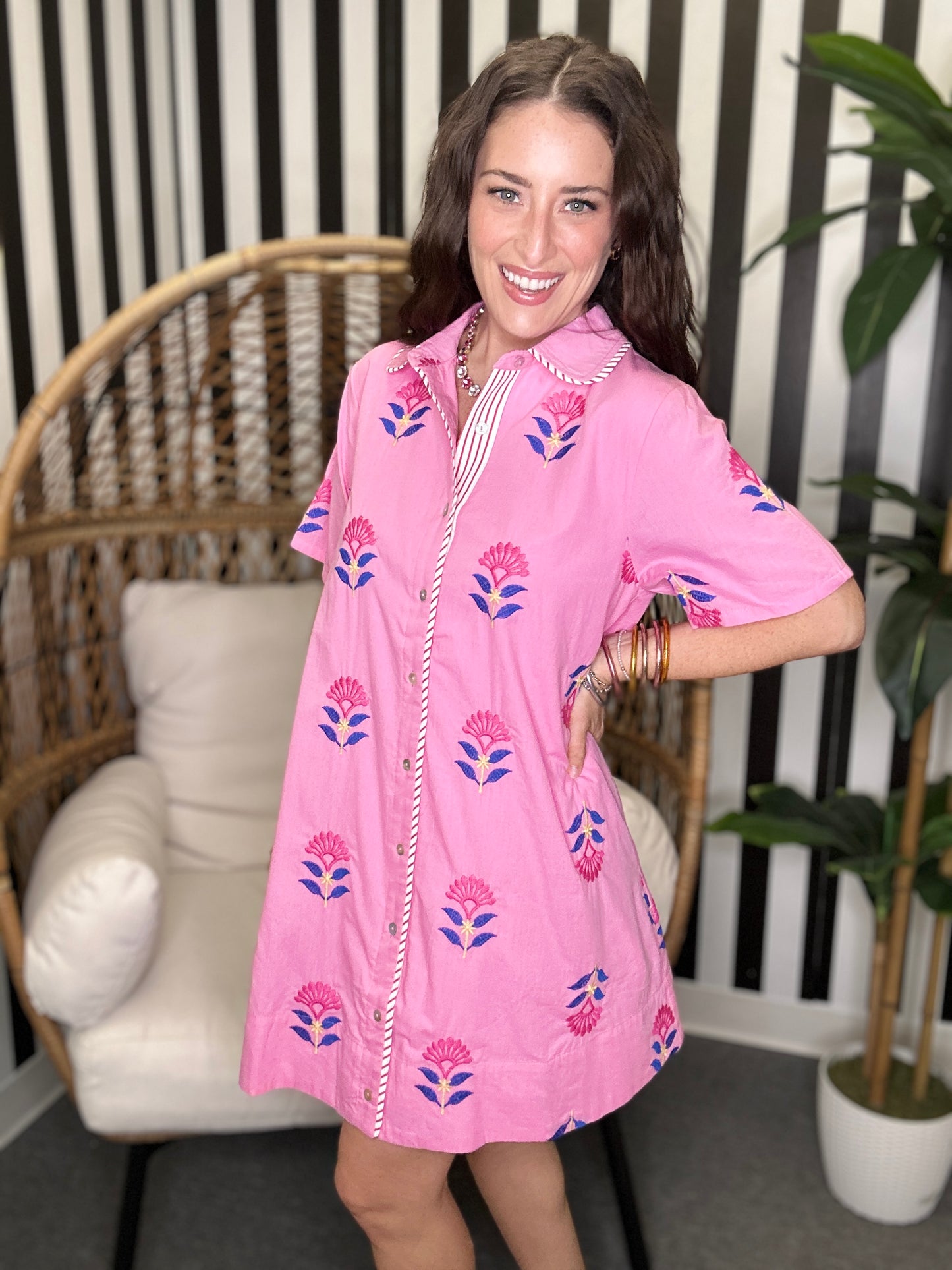 Woman wearing a pink dress with floral patterns in a room with a striped wall and wicker chair.