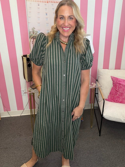 A woman wearing a green and white vertically striped midi dress with puff sleeves and a button-down front, standing in front of a pink and white striped wall.
