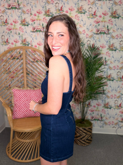 Woman in a denim dress standing in a room with floral wallpaper, wicker chair, and plant.