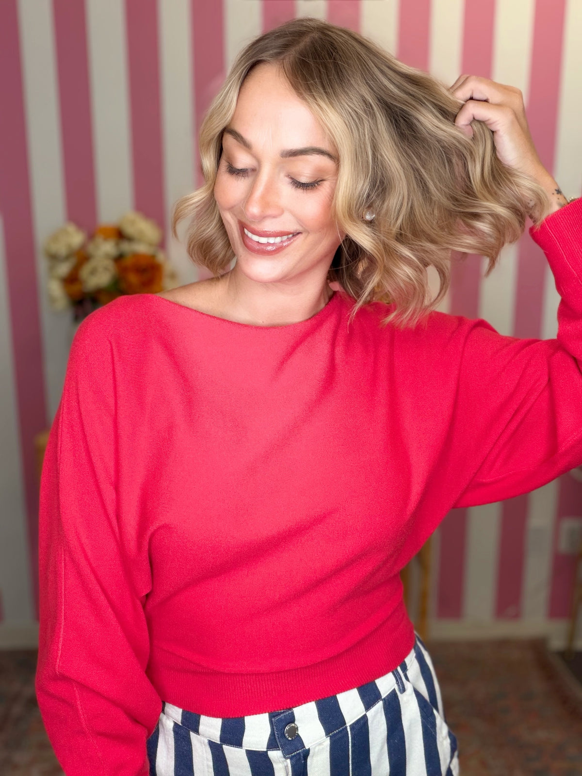 A woman with blonde wavy hair wearing a bright red long-sleeve knit top and blue-and-white striped pants, posing in front of pink and white striped wallpaper.