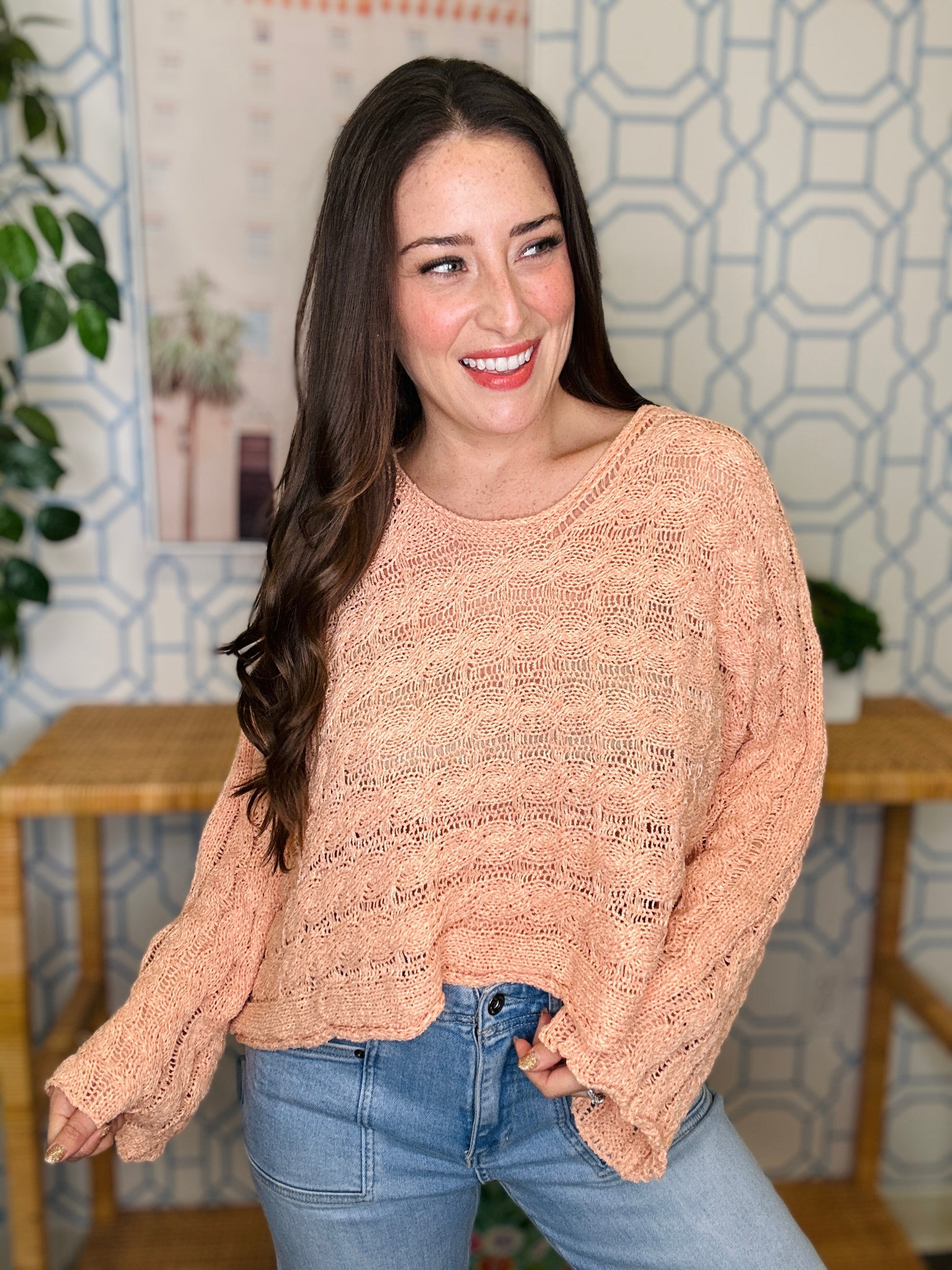 Woman wearing a beige sweater and blue jeans standing in a room with a decorative wall.