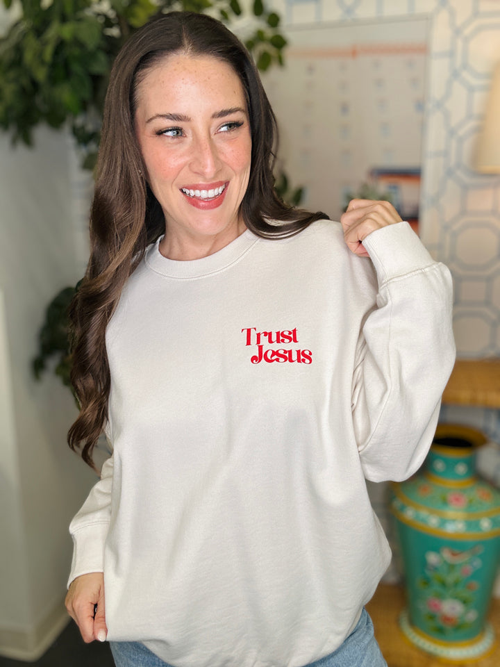 Woman wearing a white sweatshirt with 'Trust Jesus' in red text, standing indoors.