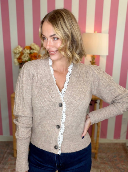 woman standing in a taupe cardigan with lace in a room with pink and white stripes.